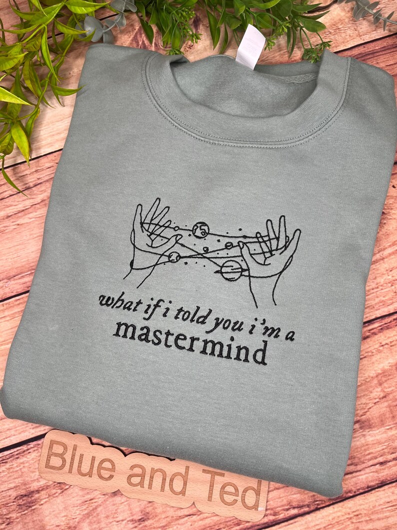 May include: A light blue sweatshirt with a black graphic of hands holding planets and stars. The text "what if i told you i'm a mastermind" is printed below the graphic.