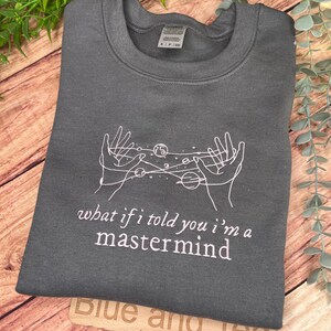May include: Gray sweatshirt with a white outline of two hands holding a galaxy with planets and stars. The text "what if i told you i'm a mastermind" is printed in pink below the galaxy.