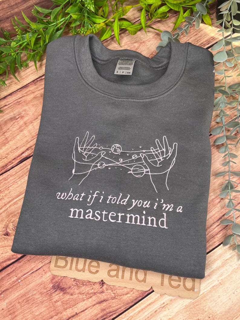 May include: Gray sweatshirt with a white outline of two hands holding a galaxy with planets and stars. The text "what if i told you i'm a mastermind" is printed in pink below the galaxy.