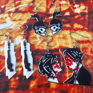 Pode incluir: Uma coleção de brincos feitos à mão com vários designs. Os brincos apresentam imagens de uma caveira com capacete, uma figura pendurada e um casal com respingos de sangue. Os brincos são pretos, brancos e vermelhos.