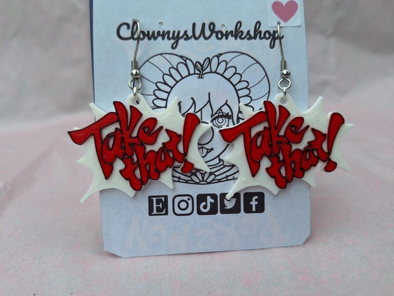 May include: A pair of white and red earrings with the words "Take That!" on each earring. The earrings are shaped like a starburst.