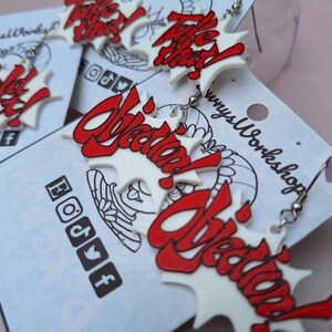 May include: A pair of white and red earrings with the word "Objection!" written in a bold, stylized font. The earrings are on a white card with the words "Clowny's Workshop" printed on it.