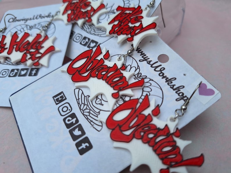 May include: A pair of white and red earrings with the word "Objection!" written in a bold, stylized font. The earrings are on a white card with the words "Clowny's Workshop" printed on it.