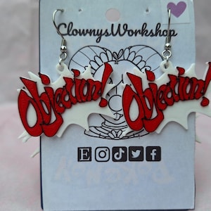 May include: A pair of white and red acrylic earrings with the word "Objection!" in red lettering. The earrings are hanging from silver hooks.