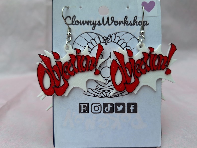 May include: A pair of white and red acrylic earrings with the word "Objection!" in red lettering. The earrings are hanging from silver hooks.