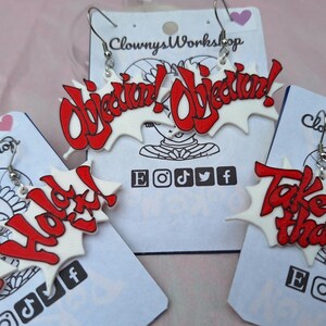 May include: Three pairs of white and red acrylic earrings with the words "Objection! Objection!" "Hold It!" and "Take That!" printed in red. The earrings are on white cards with the words "Clowny's Workshop" and social media icons.