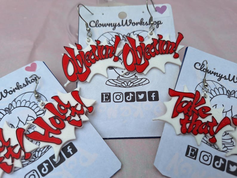 May include: Three pairs of white and red acrylic earrings with the words "Objection! Objection!" "Hold It!" and "Take That!" printed in red. The earrings are on white cards with the words "Clowny's Workshop" and social media icons.