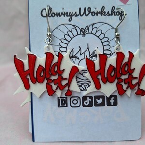 May include: A pair of white and red earrings with the words "Hold It!" on each earring. The earrings are made of acrylic and have silver hooks.
