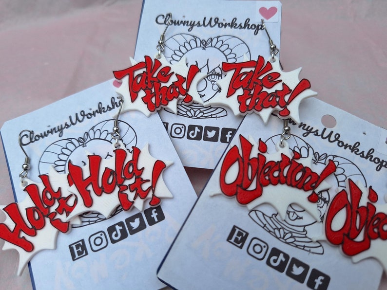 May include: A set of four pairs of earrings featuring red and white text on a white background. The earrings are shaped like a speech bubble and feature the phrases "Take That!" "Hold It!" and "Objection!"