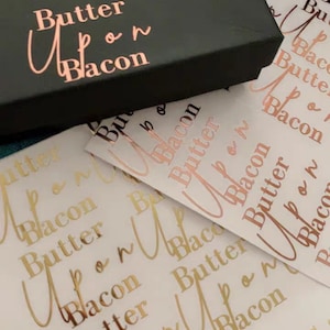 May include: A black box with the words "Butter Upon Bacon" in rose gold lettering. The box is sitting on top of a sheet of white paper with the same words printed in gold lettering.