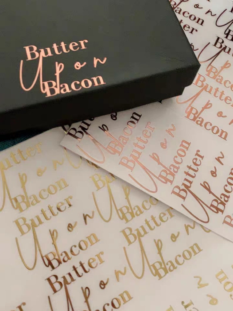 May include: A black box with the words "Butter Upon Bacon" in rose gold lettering. The box is sitting on top of a sheet of white paper with the same words printed in gold lettering.
