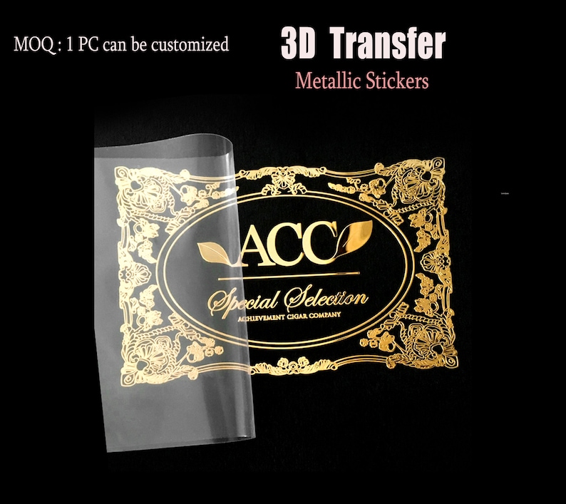 May include: Gold metallic sticker with the text "ACC Special Selection ACHIEVEMENT CIGAR COMPANY" in a decorative frame. The sticker is on a black background and is being peeled off of a clear sheet.