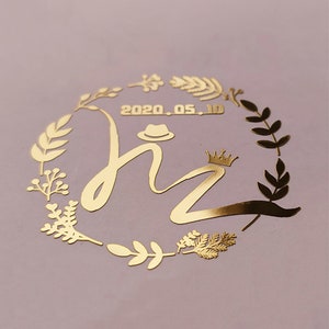 May include: Gold foil sticker with a wreath of leaves and the letters "hz" in a cursive font. The letters are topped with a crown and a hat. The date "2020.05.10" is written above the letters.