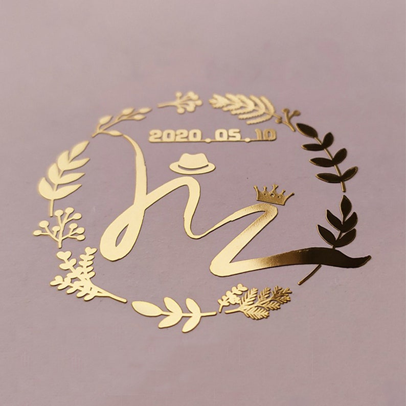 May include: Gold foil sticker with a wreath of leaves and the letters "hz" in a cursive font. The letters are topped with a crown and a hat. The date "2020.05.10" is written above the letters.