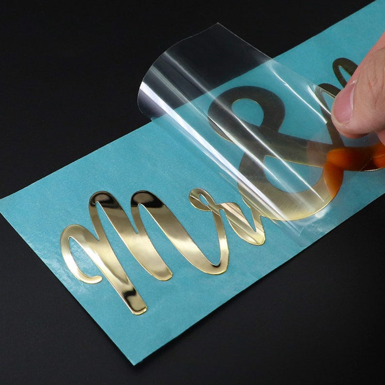 May include: Gold metallic letters spelling "Mr & Mrs" on a blue backing sheet. The letters are being peeled off the backing sheet.