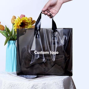 Pode incluir: Uma sacola de compras transparente com alças pretas e o texto "Custom logo". Dentro da sacola há uma peça de roupa marrom dobrada. A sacola é segurada por uma mão. A sacola está sobre uma toalha de mesa branca.