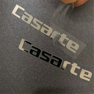 May include: Two silver metal stickers with the word "Casarte" in a modern font. The top sticker is being peeled off of a dark gray surface.