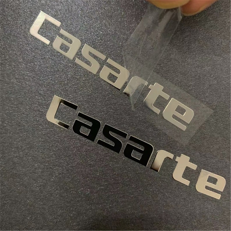 May include: Two silver metal stickers with the word "Casarte" in a modern font. The top sticker is being peeled off of a dark gray surface.