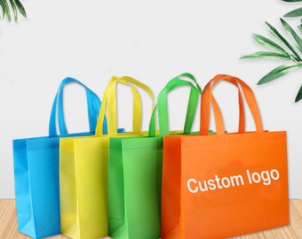 Puede incluir: Cuatro bolsas de mano coloridas en azul, amarillo, verde y naranja, cada una con asas a juego. La bolsa naranja tiene el texto "Custom logo" en blanco. Las bolsas están hechas de un material no tejido y son adecuadas para compras o promociones.