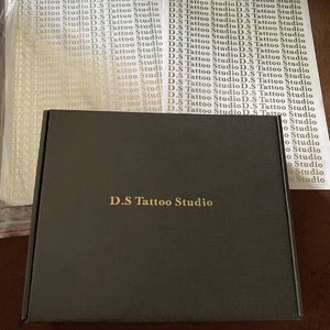 May include: A black box with the text "D.S Tattoo Studio" printed in gold on the front. The box is surrounded by sheets of white paper with the text "D.S Tattoo Studio" printed in gold.