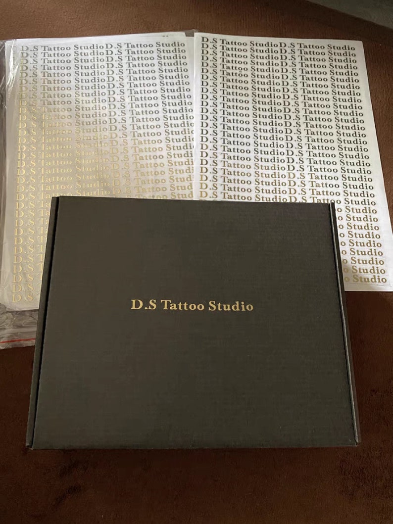 May include: A black box with the text "D.S Tattoo Studio" printed in gold on the front. The box is surrounded by sheets of white paper with the text "D.S Tattoo Studio" printed in gold.