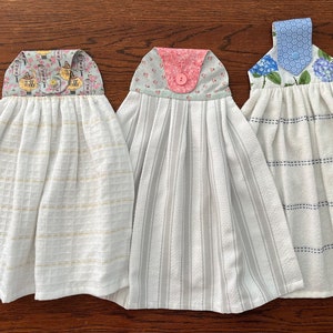 May include: Three white kitchen towels with different colored and patterned fabric accents. The towels are hanging on a wooden surface.