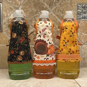May include: Three clear plastic bottles with orange, green, and yellow liquid inside. Each bottle is covered with a fabric apron with a different Thanksgiving-themed pattern. The aprons are tied with orange ribbon.