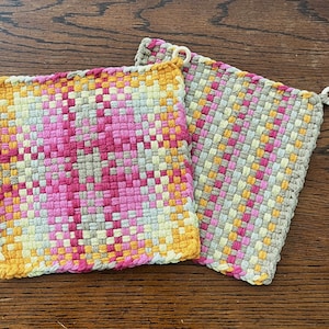 May include: Two woven pot holders with a pink, yellow, and gray geometric pattern. The pot holders have a white loop on each corner.