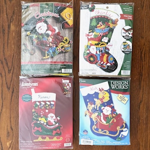 May include: Four Christmas stocking felt kits. The kits feature Santa Claus, reindeer, and other holiday designs. The stockings are in various colors, including red, green, and blue. Each kit is packaged in a clear plastic bag.