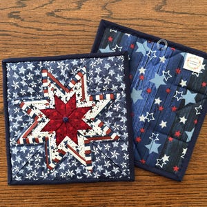 May include: Two square quilted pot holders. One features a red and white starburst design on a blue background with white stars. The other has a blue wood grain pattern with white, red, and blue stars. Both have a dark blue border.