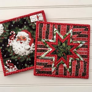May include: Two square quilted potholders with red binding. One features a vintage Santa Claus print on a black background with holly. The other displays a geometric star pattern in red, green, and white stripes with a central pine branch motif.