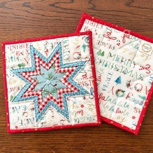 May include: Two square quilted fabric potholders with a red border. One features a central blue and red gingham star pattern on a white background with Christmas text. The other shows a white fabric with repeating holiday text and icons.