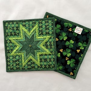 May include: Two square quilted pot holders. One features a large green star design with a button center. The other has a black background with green and gold shamrocks. Both have green borders and are likely handmade.