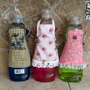 May include: Three clear plastic bottles containing different coloured liquids. Each bottle is covered with a fabric apron featuring a different pattern. The aprons have lace trim and buttons.