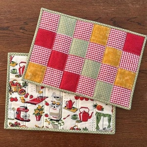 May include: Two quilted placemats, one with a red, green, and yellow checkered pattern and the other with a white background and a vintage kitchen print.