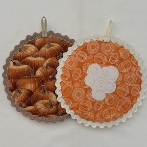 May include: Two round pot holders with scalloped edges. One pot holder features a brown and orange pumpkin print. The other pot holder features a white and orange sunflower print with a white circle in the center.