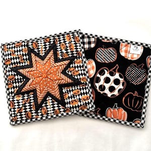 May include: Two square quilted pot holders. One features a starburst design in orange and black, with a black and white checkered border. The other has a black background with various pumpkin patterns in orange and white, and a black and white checkered border.