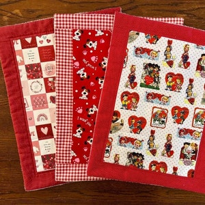 May include: Three red and white checked fabric placemats with a red border. The centre of each placemat features a different design. One placemat has a red and white polka dot background with a repeating pattern of vintage Valentine's Day cards. Another placemat has a red background with a repeating pattern of white paw prints and the words "I woof you". The third placemat has a red and white gingham pattern.