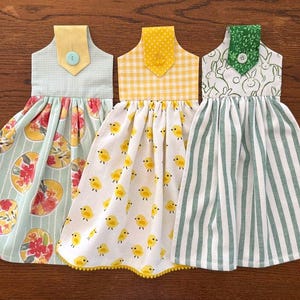 May include: Three decorative kitchen hand towels in the shape of dresses. The towels feature various patterns, including floral eggs, yellow chicks, and green stripes. Each towel has a different colored top with a button detail.
