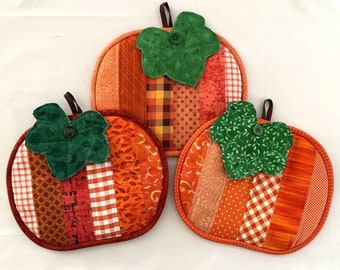 Op de afbeelding: Drie handgemaakte pannenlappen in de vorm van een pompoen. Elke pannenlap heeft een patchwork-ontwerp in tinten oranje, rood en bruin, met een groen blad en een knoopdetail. Er is een bruine lus bevestigd om op te hangen.