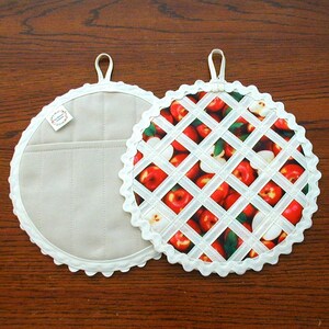 May include: Two round pot holders. One is solid beige with a pocket and a scalloped edge. The other has a white lattice design over a red apple print, also with a scalloped edge. Both have a loop for hanging.
