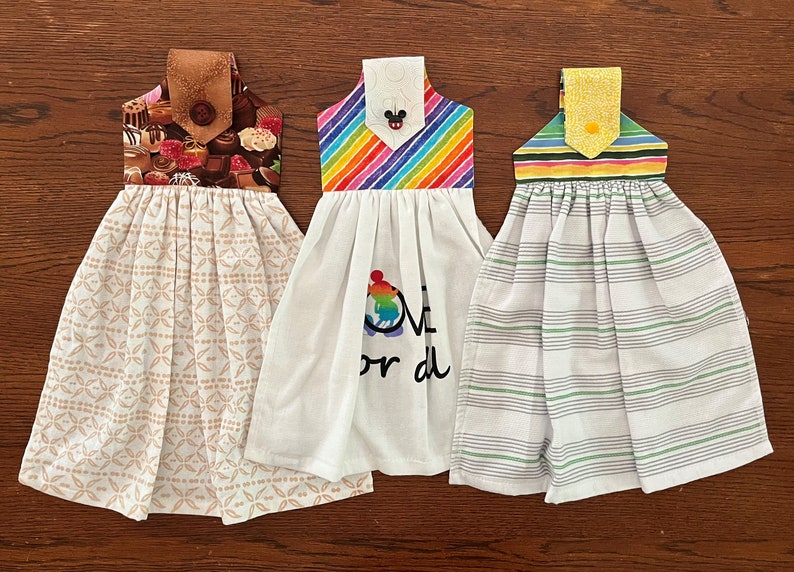 May include: Three kitchen towels with different designs. The first towel has a brown and white pattern with a chocolate design on the top. The second towel has a rainbow design on the top and the words "Love or die" printed on the bottom. The third towel has a green and white striped design with a yellow and green striped design on the top.