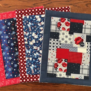 May include: Three red, white and blue fabric placemats with different patterns. The placemats have a red border with white stars, a red and white polka dot border, and a blue border with a subtle texture.