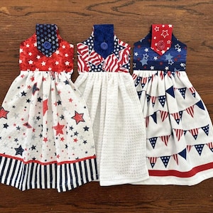 May include: Three kitchen towels with a patriotic theme. The towels are white with red, white, and blue stars, flags, and bunting patterns. Each towel has a blue button and a loop for hanging.