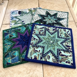 May include: Four square quilted pot holders with starburst designs. The pot holders feature various shades of blue, green, and purple fabrics. Each pot holder has a different pattern and color scheme, with a button in the center.
