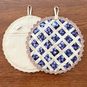May include: Two round pot holders. One is beige with a pocket and a loop for hanging. The other is a blueberry pie design with a lattice crust and a scalloped edge. The pie filling is a dark blue and purple pattern.