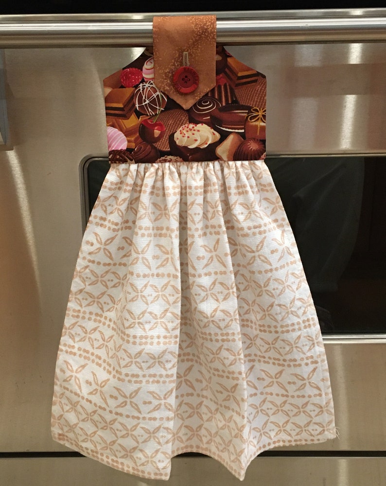May include: A white kitchen towel with a brown geometric pattern. The towel has a brown fabric loop with a red button and a chocolate-themed print.