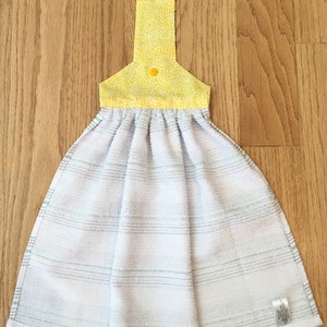 May include: A white kitchen towel with green stripes and a yellow floral print hanging loop. The loop has two yellow buttons.