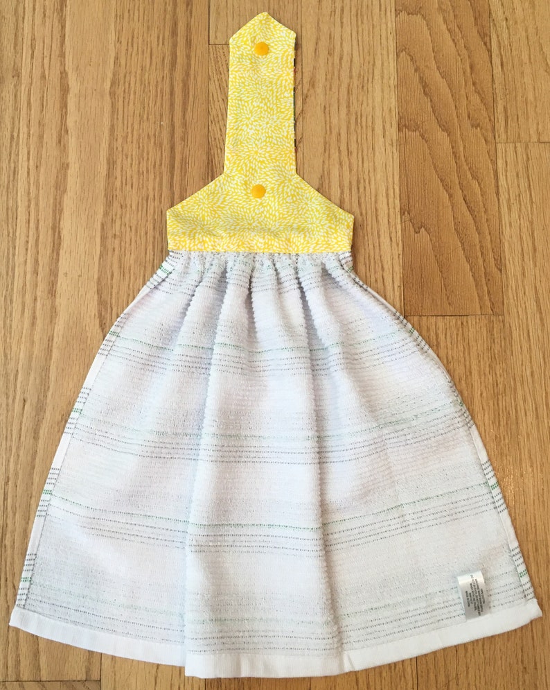 May include: A white kitchen towel with green stripes and a yellow floral print hanging loop. The loop has two yellow buttons.