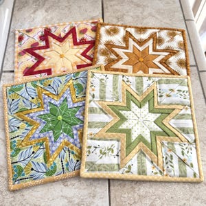 May include: Four handmade quilted star-shaped pot holders in various colors and patterns. Each square pot holder features a central star design with contrasting fabrics, including floral, striped, and sunburst patterns. The edges are finished with a coordinating binding.